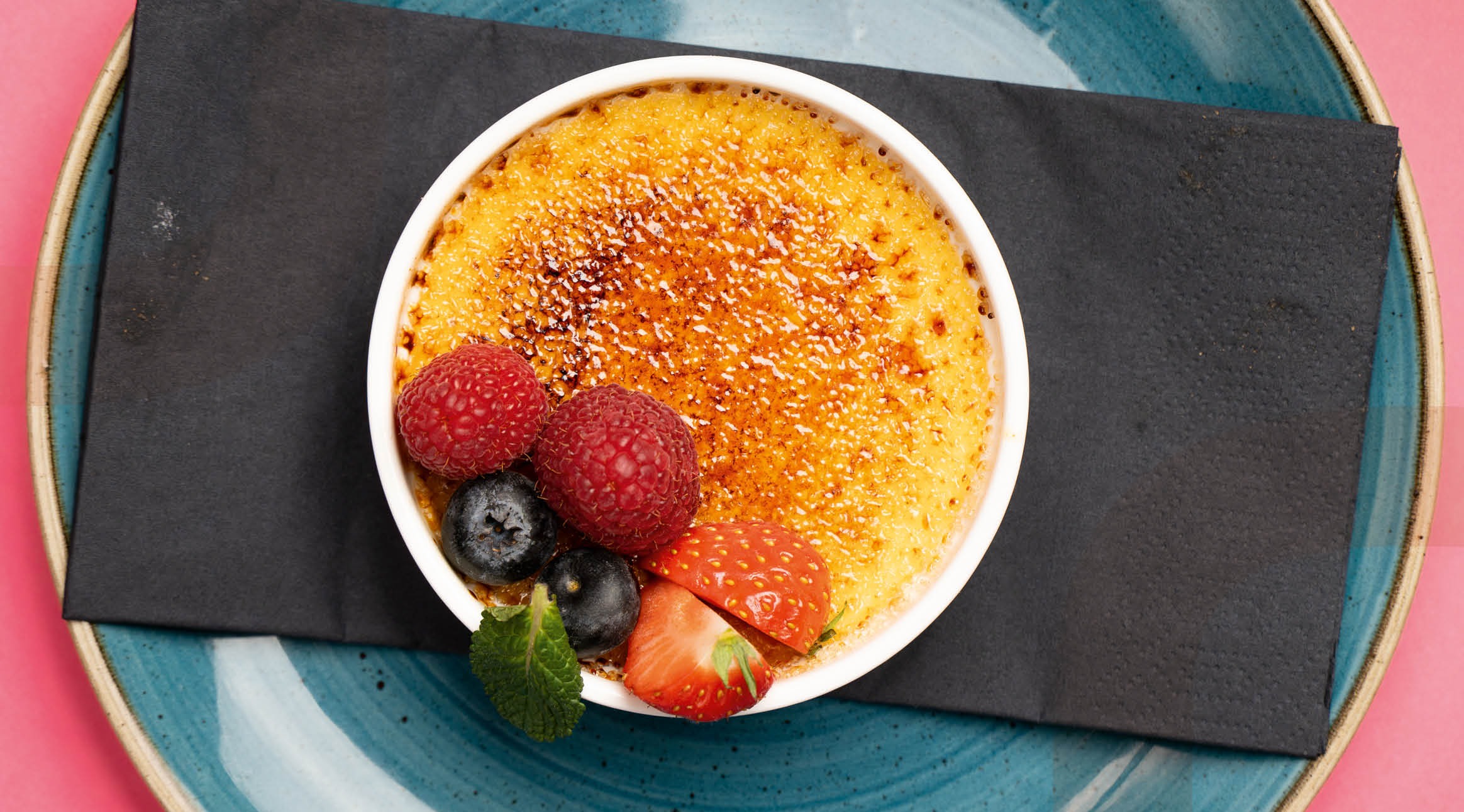 Crème brûlée in a small round ramekin garnished with raspberries, blueberries, strawberry pieces and a mint leaf. Served on a turquoise plate.