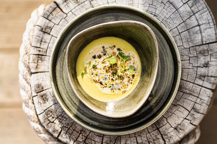 Ceramic bowl of yellow soup garnished with microgreens and spices, served on a wooden stump.