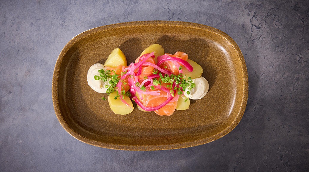 Ruskealla keraamisella vadilla tarjoiltu annos lohta, perunoita, kastiketta, pinkkiä sipulia ja vihreitä yrttejä harmaata taustaa vasten.