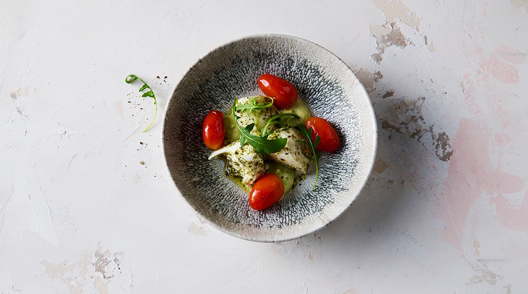 Fish in green sauce with cherry tomatoes and rocket in a grey patterned bowl on a light surface.
