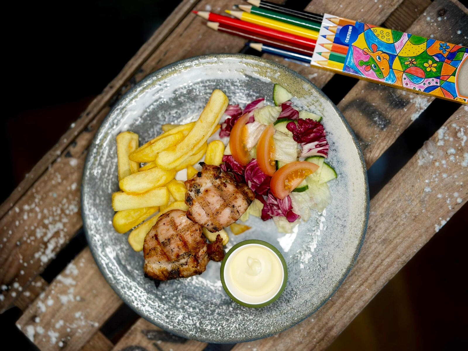 A meal of grilled chicken, french fries, salad and mayonnaise dip on a ceramic plate. Colored pencils rest nearby on the wooden table.