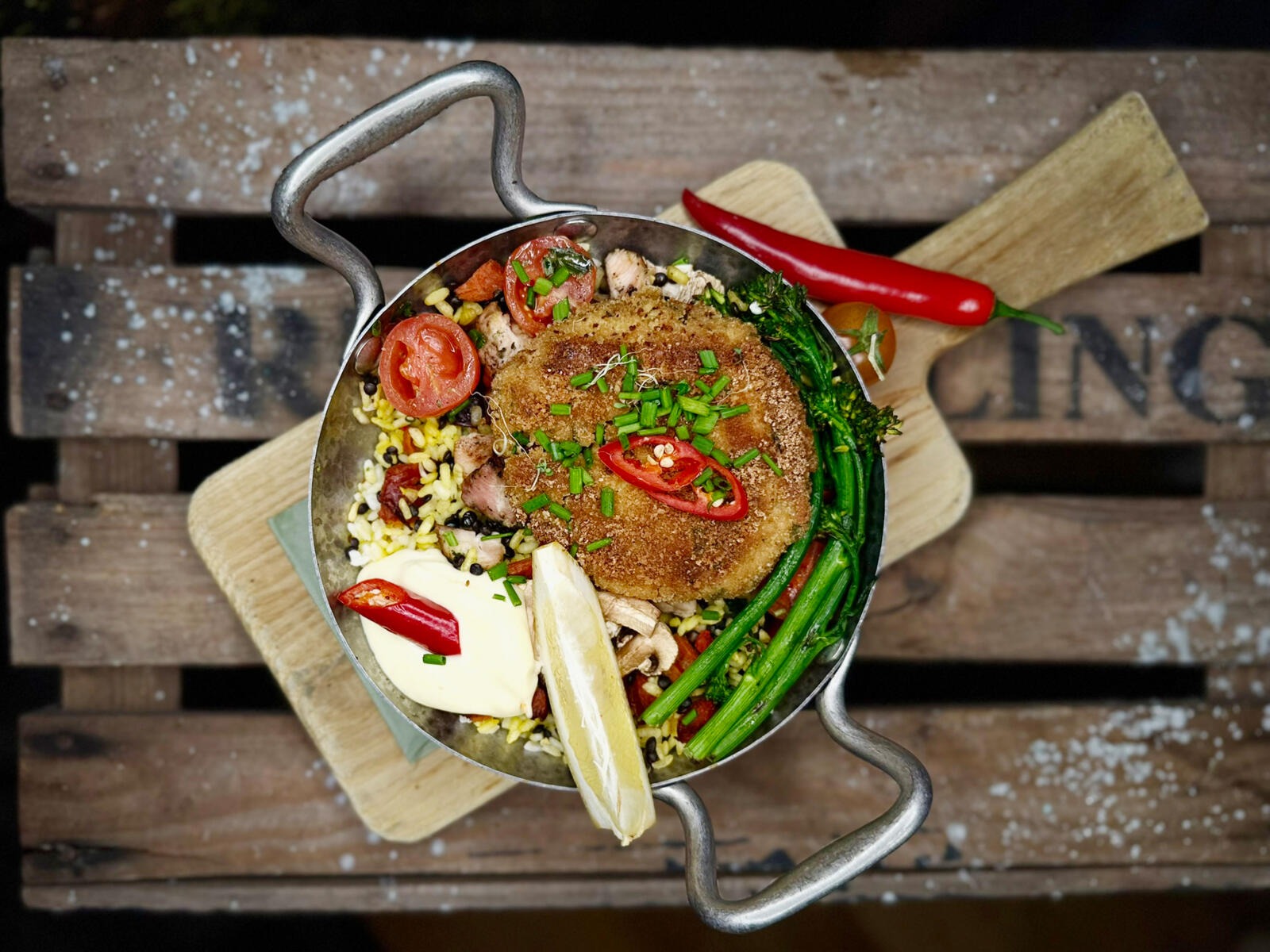 Metal pan meal with breaded cutlet, rice, broccolini, tomatoes, green onions and red chili on a wooden board.