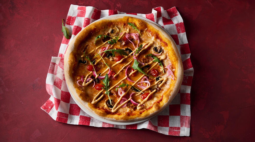 Pizza on a red table, placed on a checkered cloth. Toppings include mushrooms, cherry tomatoes, red onion, rocket and drizzled sauce.