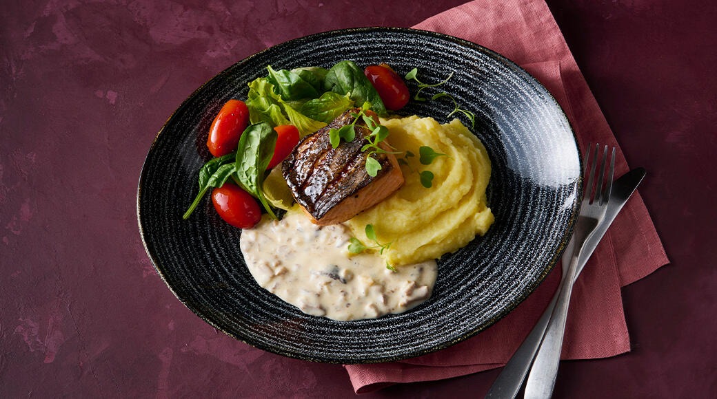 Grilled salmon with mashed potatoes, creamy mushroom sauce, salad and cherry tomatoes on a black plate. A fork and knife lie on a red napkin.