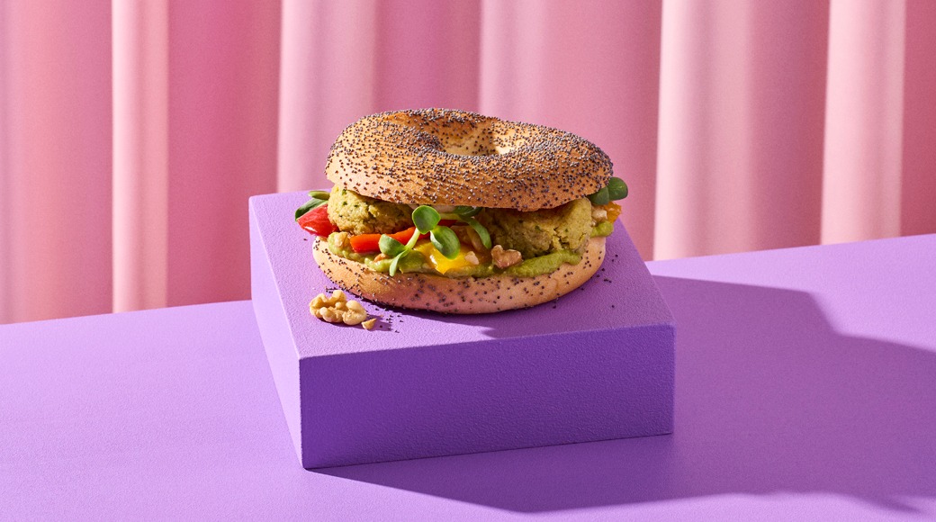 Unikoilla ripoteltu bagel, jonka välissä on vihreää levitettä, kasvispihvi, paprikaa ja versoja. Bagel on violetin korokkeen päällä vaaleanpunaista verhoa vasten.
