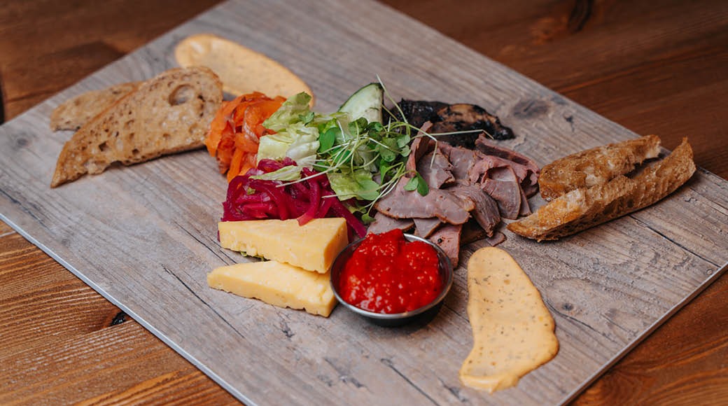 Lautanen leikkeleitä, juustoa, leipää ja salaattia puisella tarjoilualustalla. Keskellä pieni kulho punaista kastiketta.
