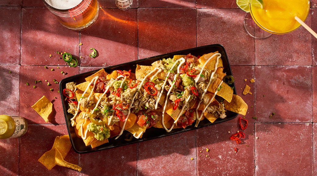 Alus nachosid, millel on tõmmatud liha, guacamole, tšilli ja kreemkastmeribad. Kõrval on õlu ja kollane kokteil.