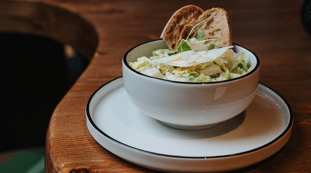 Valkoinen kulho caesar-salaattia, jossa parmesanlastuja ja leivänpaloja, tarjoiltuna puisella pöydällä valkoisella lautasella.