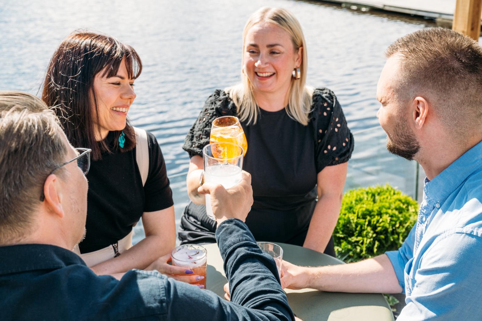 Cheerful group enjoying drinks in sunshine at a table by the water, raising glasses in a toast.