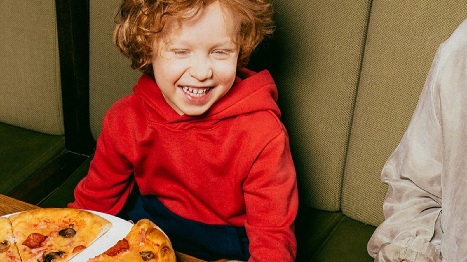 Naurava lapsi istuu pöydän ääressä vihreässä verhoillussa penkissä ja pizzaa lautasella