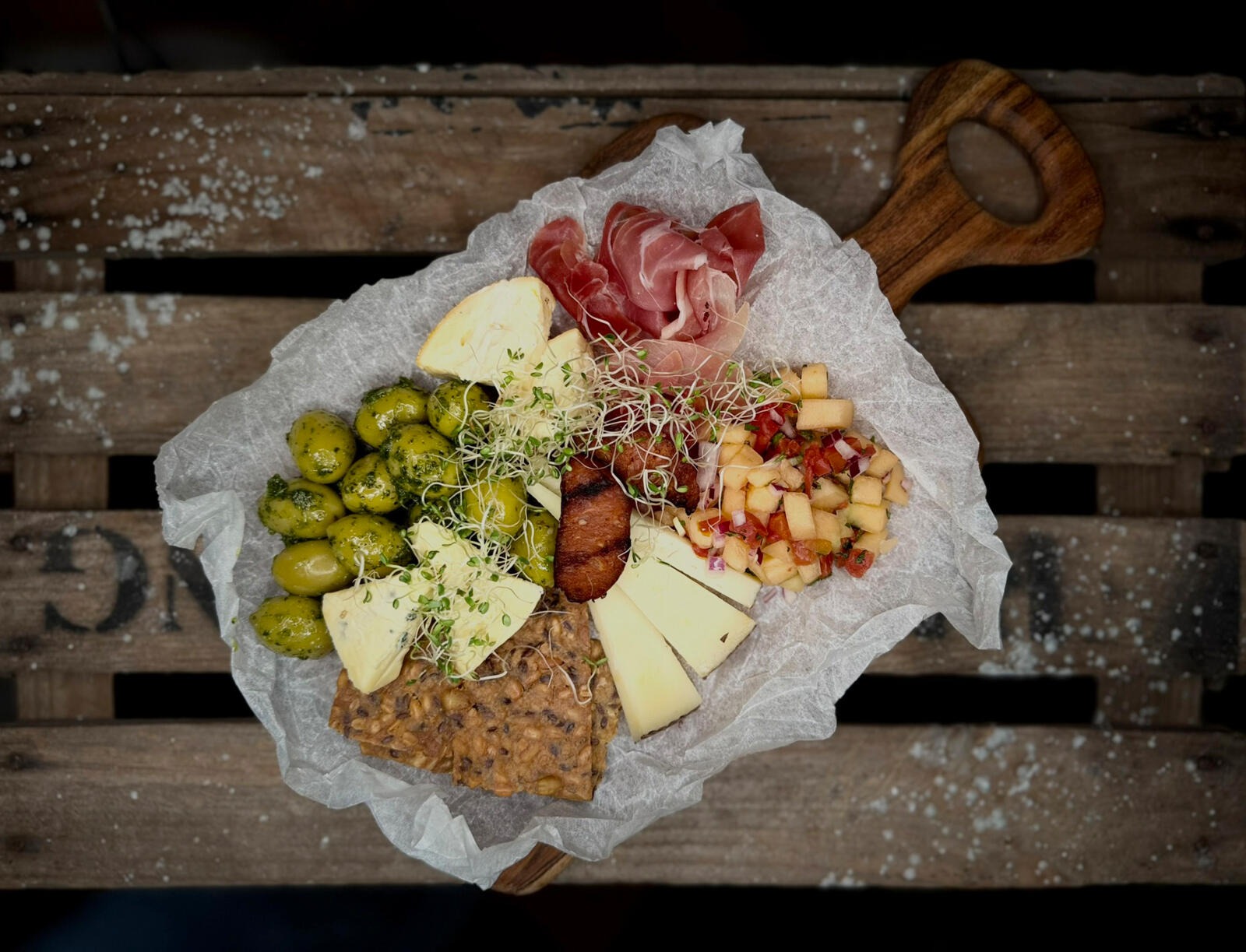 Charcuterie board on wooden platter featuring olives, prosciutto, cheese slices, seeded crackers, and fruit salsa garnished with sprouts.