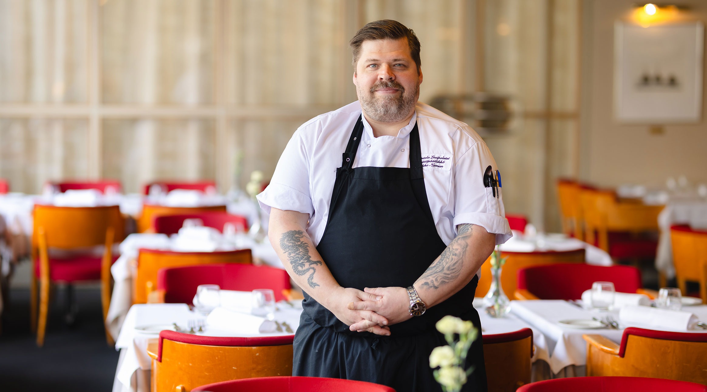 Chef stands in a restaurant dining room with set tables and red upholstered chairs. The space is lit with natural light.