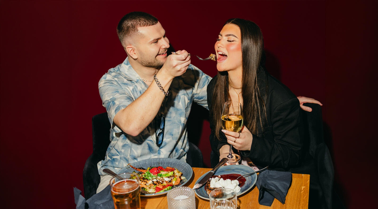 Ett par äter middag i en restaurang med röda väggar. En person matar den andra som håller ett vinglas. På bordet finns två maträtter och drycker.