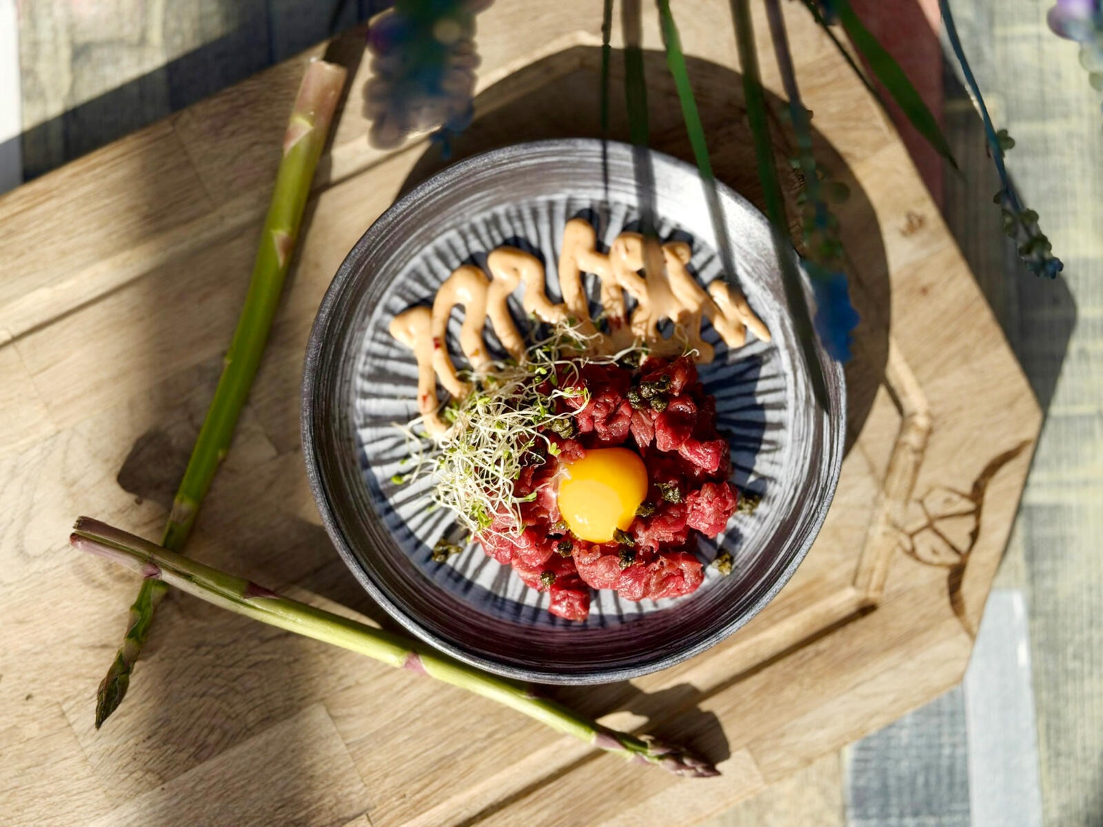 Bowl containing a tartare dish with raw meat, egg yolk, and sprouts. The bowl sits on a wooden board with asparagus.