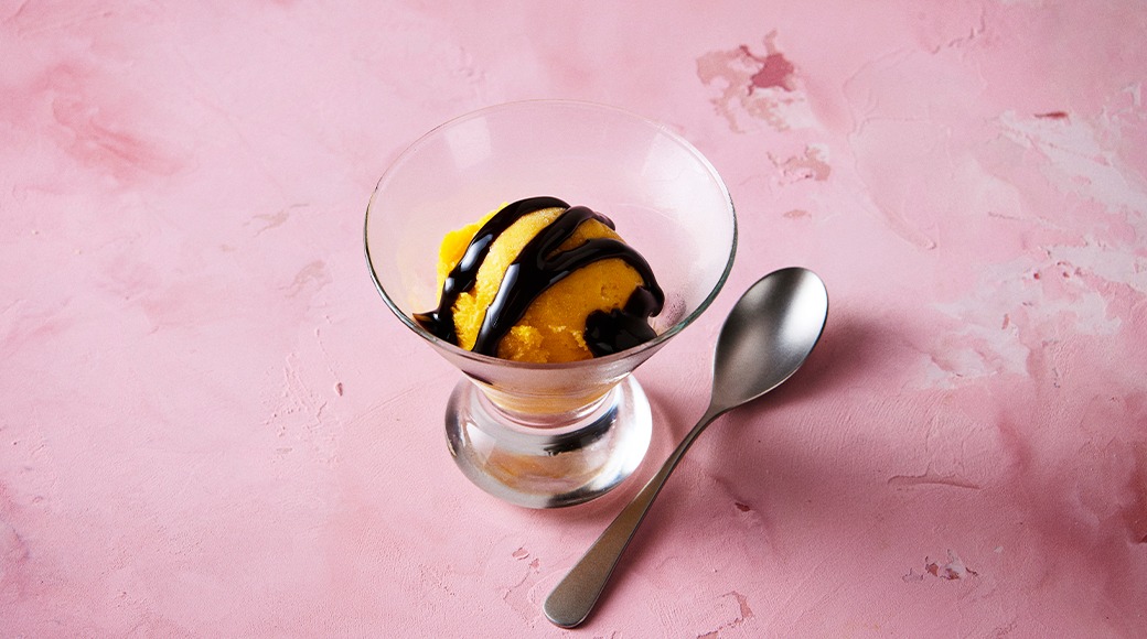 Yellow ice cream or sorbet with chocolate sauce in a glass dish. A spoon lies beside it on a pink surface.