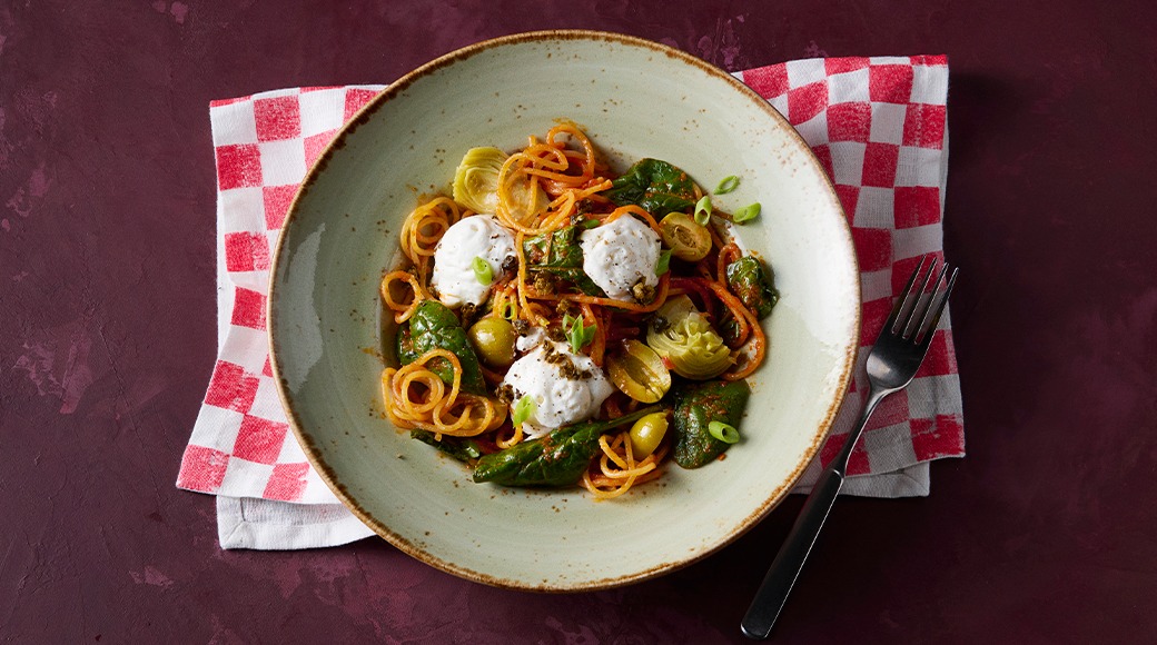 Green ceramic bowl with carrot spaghetti, spinach, artichoke, olives and mozzarella balls. The bowl rests on a red and white checkered cloth, with a fork beside it.