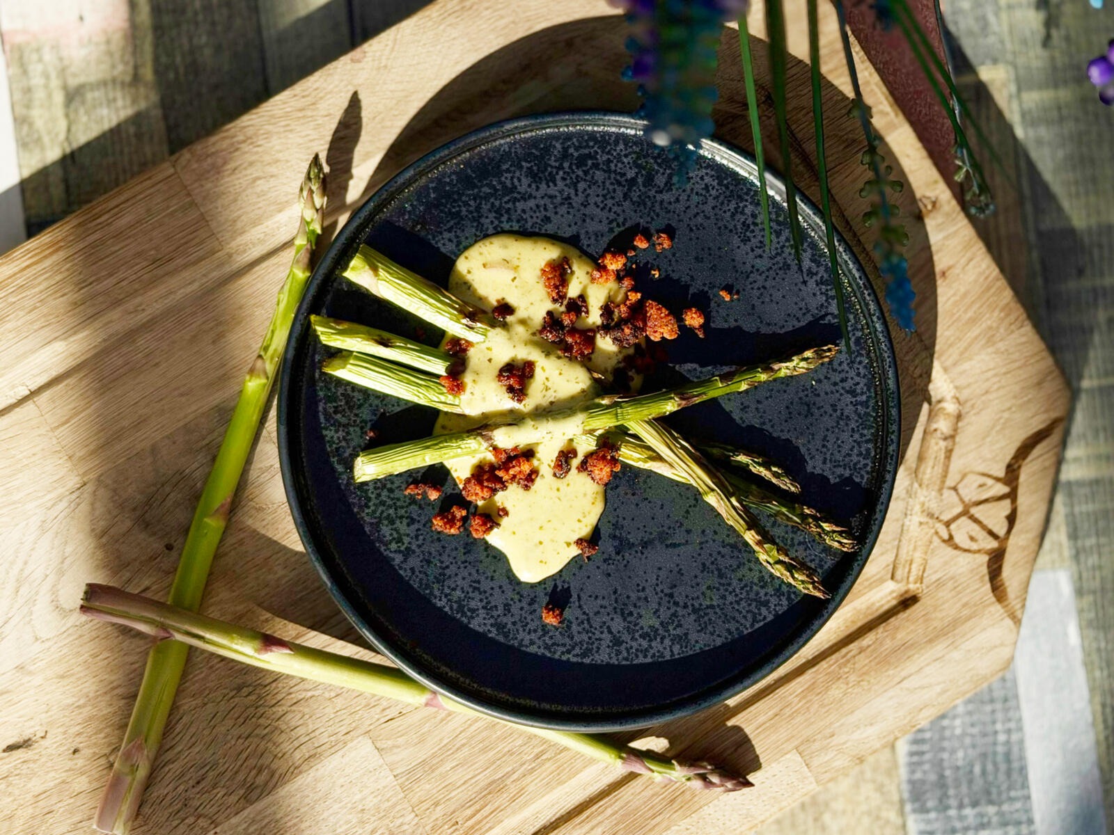 Grilled asparagus spears on a dark plate with yellow sauce and red spice crumbles on a wooden serving board.