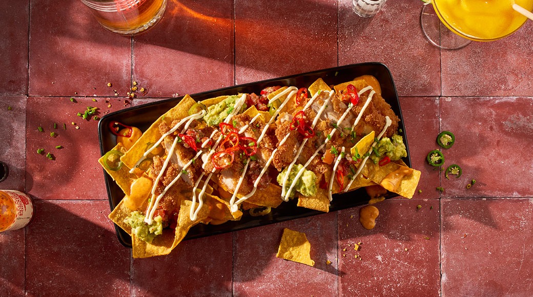 Mustal kandikul nachod krõbeda kana, guakamole, kreemja kastme, tšillilõikude ja jalapeñodega. Kõrval on joogid punasel plaaditud laual.