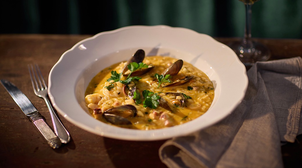 Mussel- och skaldjursrisotto i en vit tallrik på ett träbord. Bestick och ett vinglas bredvid tallriken.