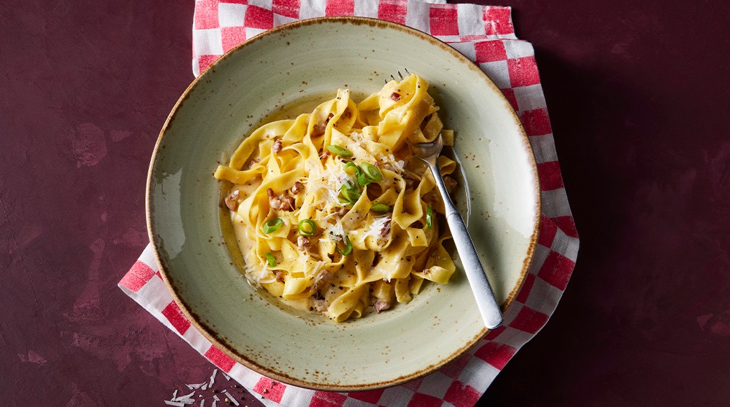 Tagliatelle in a creamy sauce topped with bacon, grated cheese and green spring onion. The dish is in a green bowl on a red and white checkered cloth.