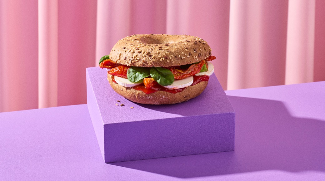 Siemenillä koristeltu täysjyväbagel, jonka välissä on mozzarellaa, pinaatinlehtiä ja aurinkokuivattuja tomaatteja. Bagel on violetin korokkeen päällä vaaleanpunaista verhoa vasten.