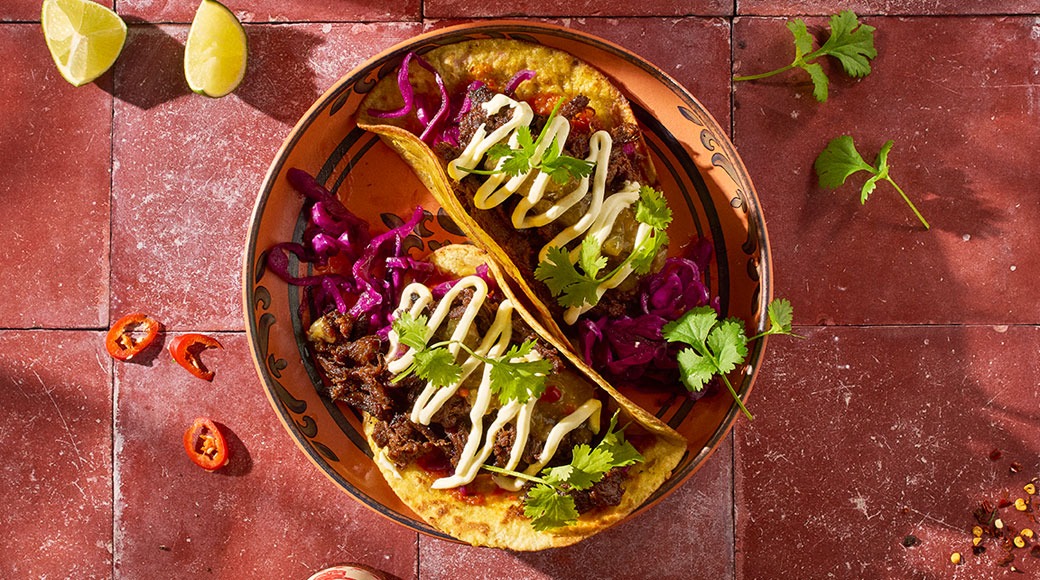 Kaksi tacoa koristellulla lautasella punaisilla laatoilla. Täytteenä lihaa, punakaalia, kastiketta ja korianteria. Ympärillä limetinpaloja ja chilirenkaita.