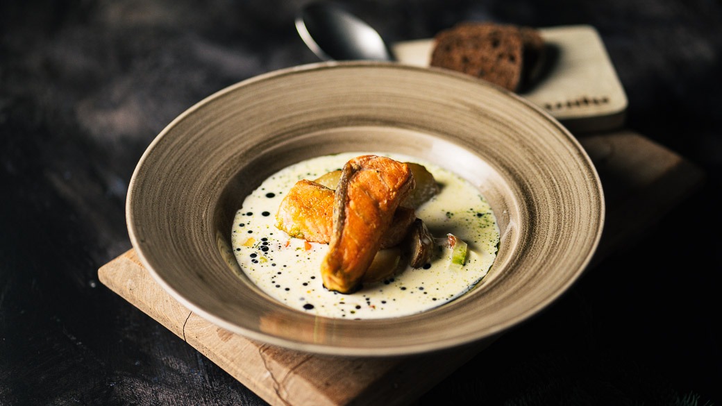 Keramiktallrik med fiskrätt i ljus soppa. Tallriken är på ett träunderlägg. En sked och bröd syns bredvid.