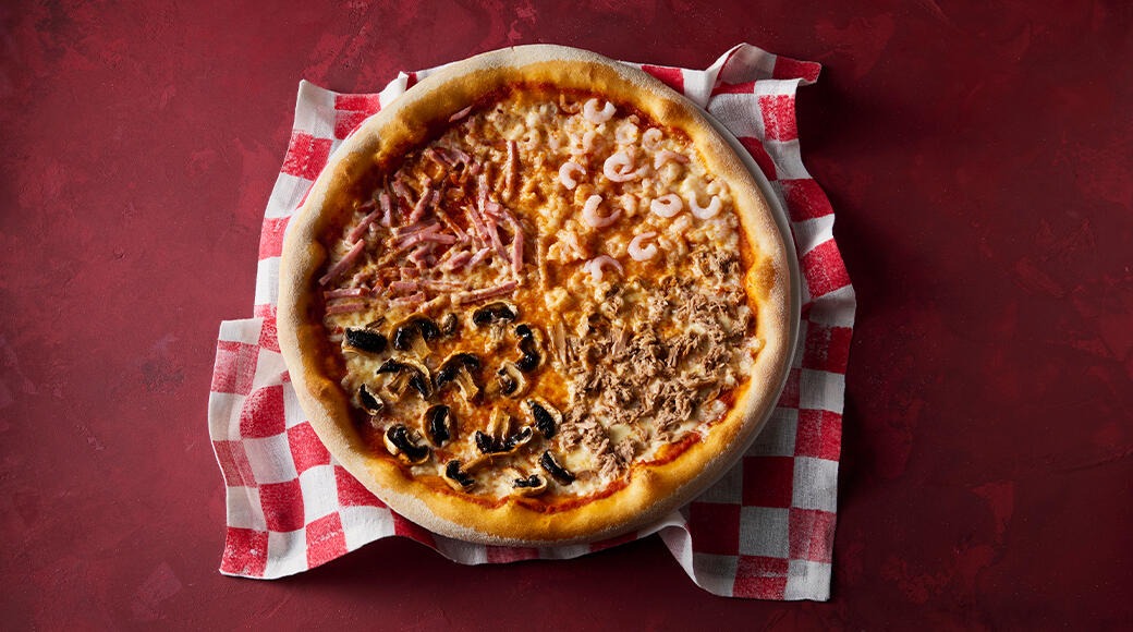 Pizza divided into four sections with ham, prawns, mushrooms and tuna. The pizza sits on a red and white checkered cloth on a red table.