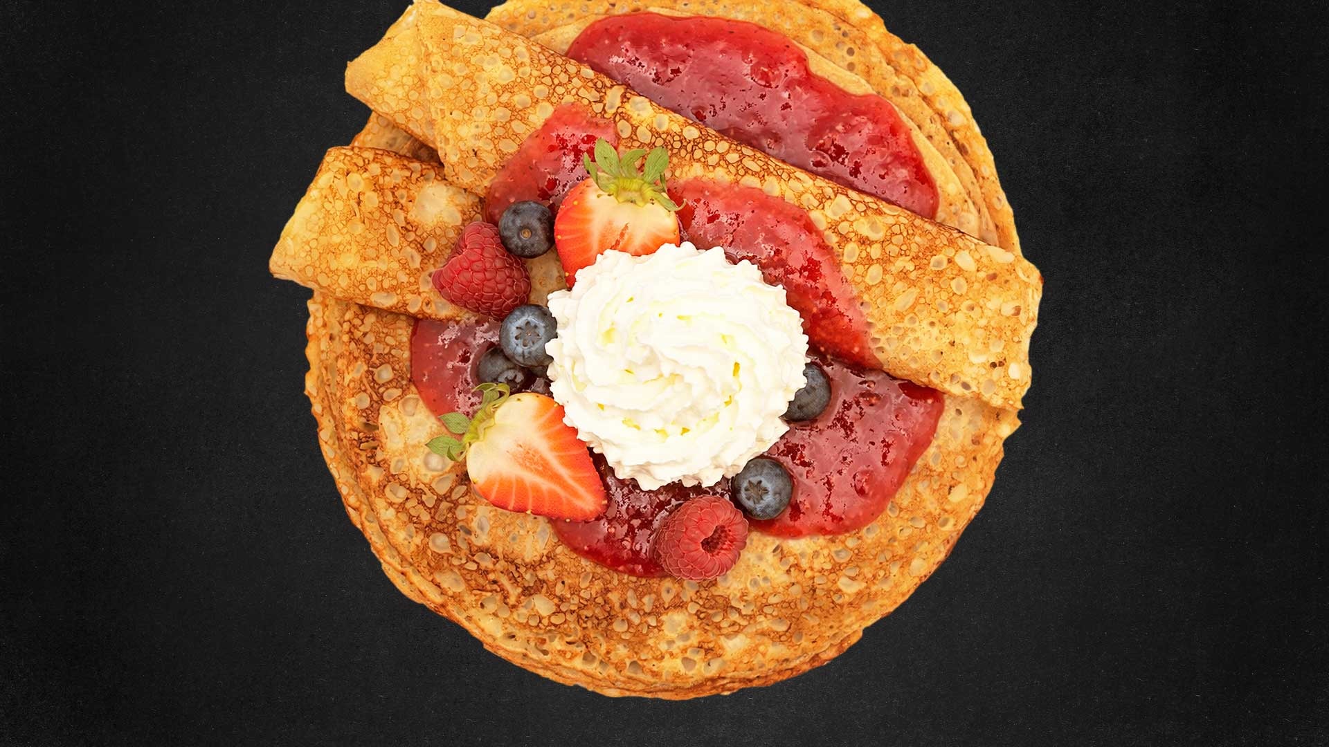 Crepe topped with red jam, whipped cream, strawberries, blueberries, and raspberries on a black background.