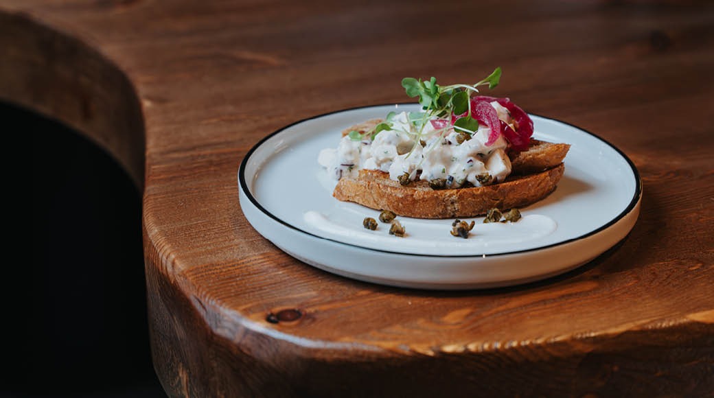 Voileipä, jossa valkoista täytettä, kaprista ja punaisia mikroyrttejä, tarjoiltuna valkoisella lautasella puisella pöydällä.