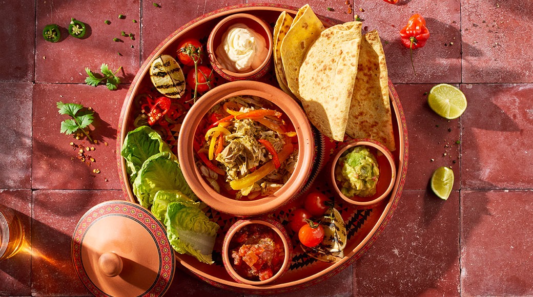 Terrakottalautasella tortillalastuja, pulled-lihaa paprikoiden kanssa, salsaa, guacamolea, smetanaa, salaattia ja tomaatteja punaisella laatalla.