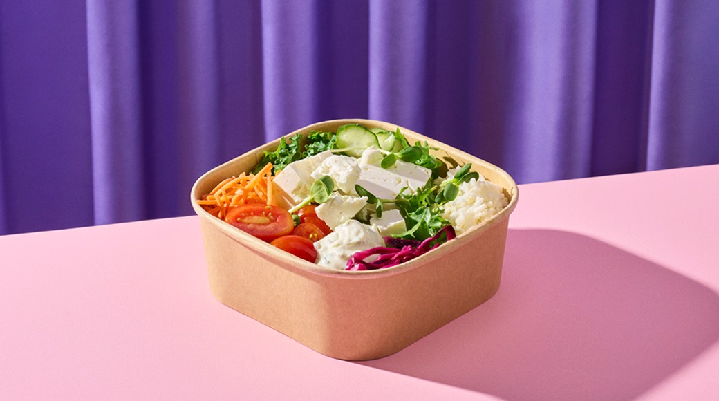 Kartonkirasiassa salaattia, jossa on fetaa, riisiä, tomaattia, porkkanaraastetta, kurkkua, lehtikaalia ja punakaalia. Rasia on vaaleanpunaisella pöydällä violetin verhon edessä.
