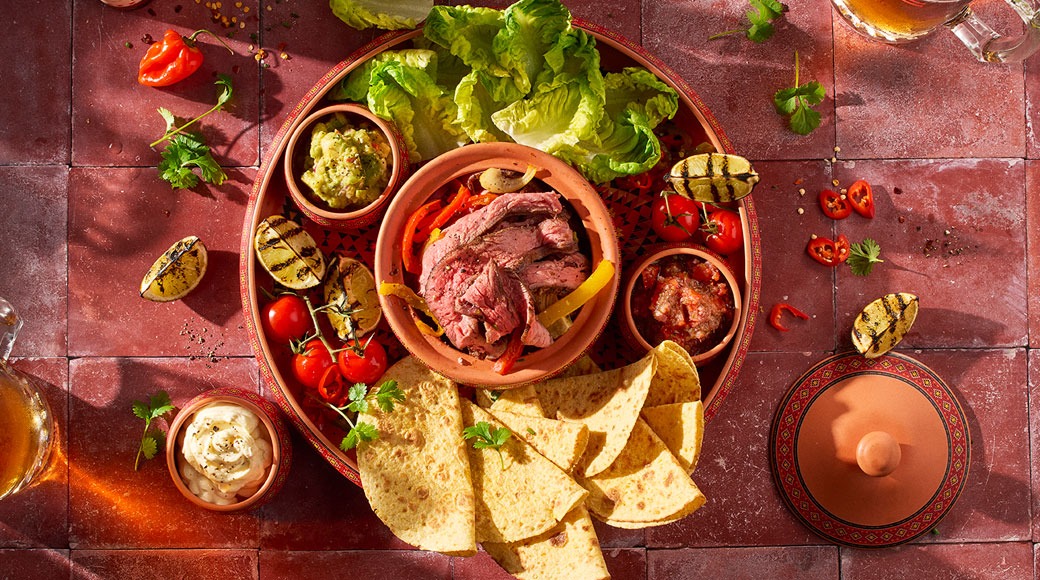 Fajita-tarjotin ylhäältä kuvattuna: savikulhossa viipaloitua grillattua lihaa ja paprikaa, ympärillä tortillakolmioita, guacamolea, salsaa, smetanaa, salaattia, tomaatteja ja grillattuja limetinpuolikkaita.