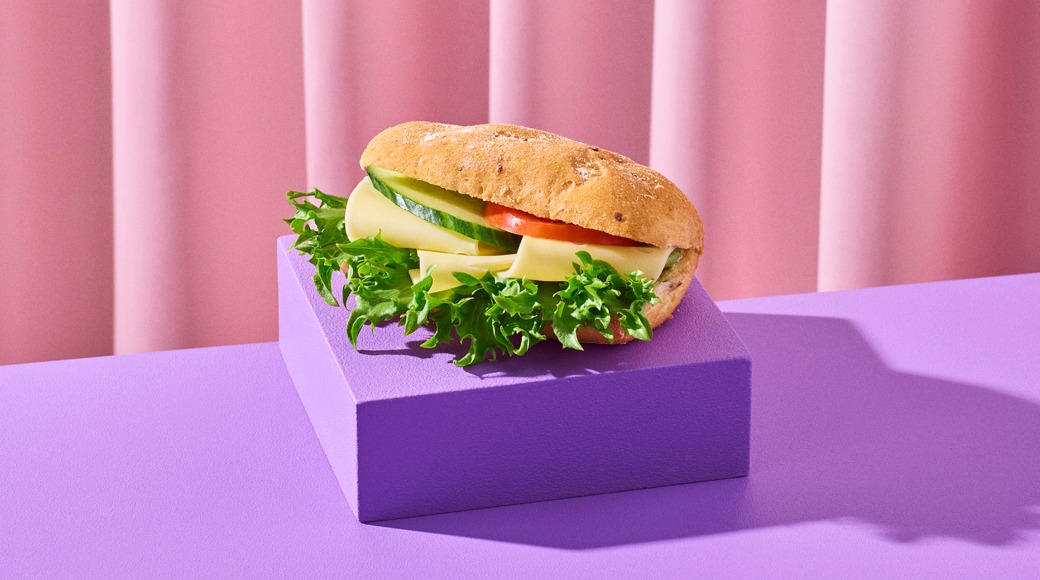 Juustosämpylä, jossa on salaattia, kurkkua ja tomaattia, violetin korokkeen päällä vaaleanpunaista verhoa vasten.