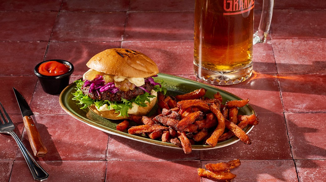 Burger ja bataadifriikartulid rohelisel taldrikul punasel plaatlaual. Kõrval on õllekruus, ketšup ning kahvel ja nuga.