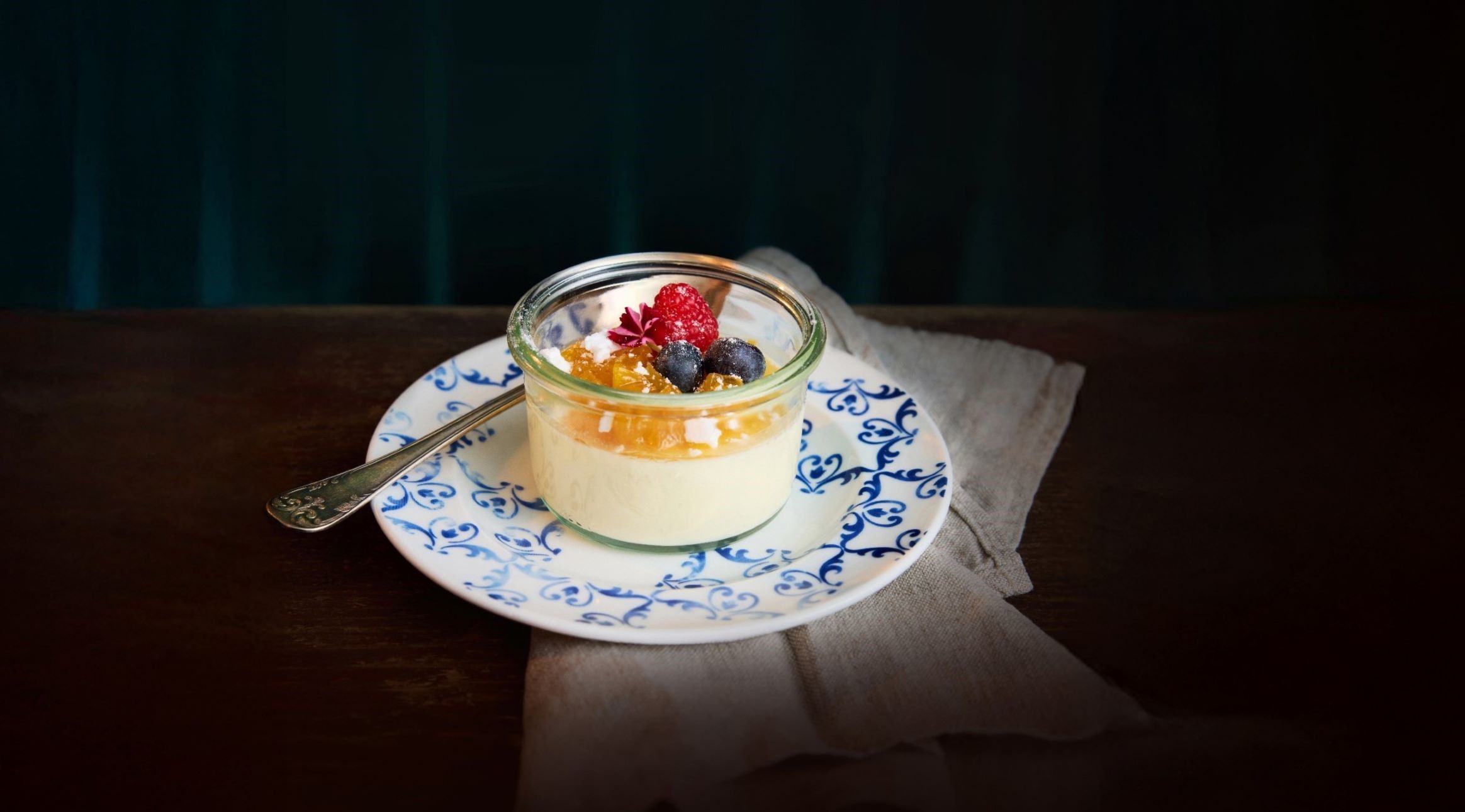 Pannacotta i en jar toppad med bär och honung på en dekorativ blå-vit tallrik.