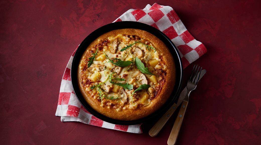 Pizza on a black pan resting on a red and white checkered cloth. It is topped with cheese, chicken, pineapple and arugula. A fork and knife lie beside it.