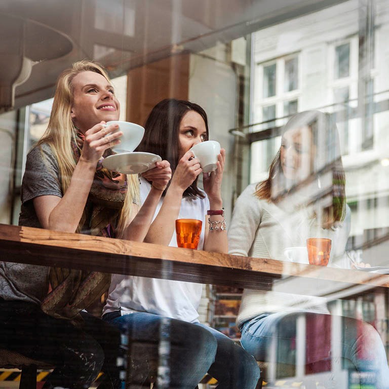 Kolme henkilöä kahvilassa ikkunan äärellä nauttimassa juomia, heijastus näkyy lasissa.