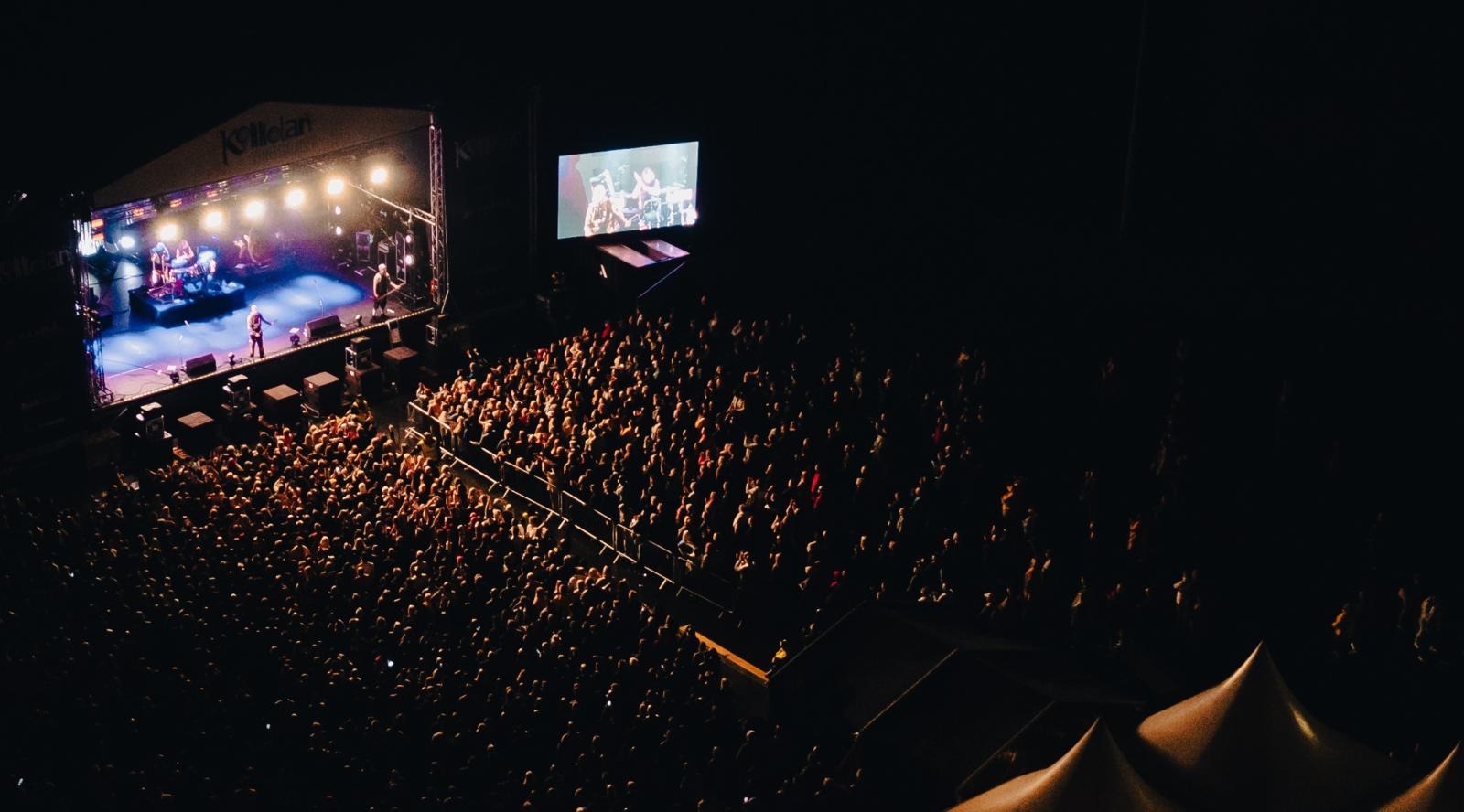 Large audience at a concert with illuminated stage on the left and musicians performing. A screen displays the performance on the right.
