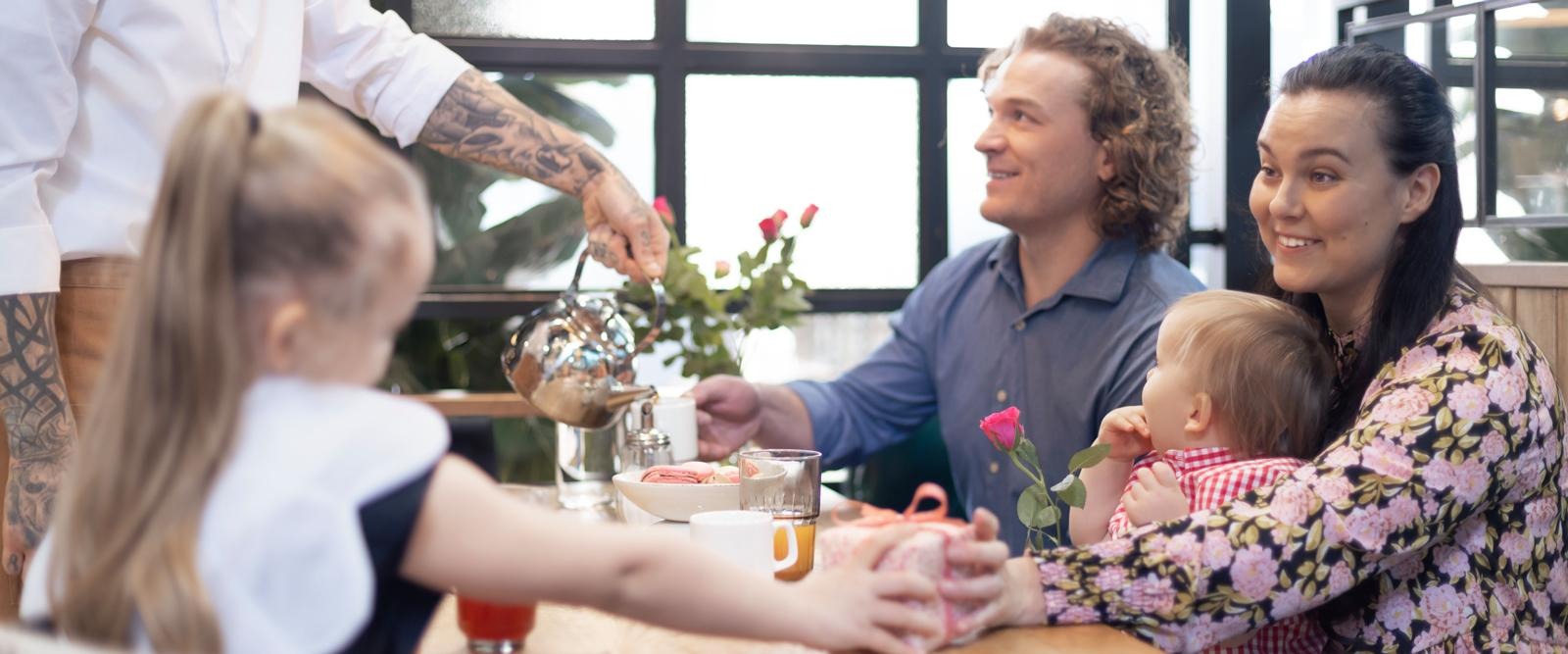 Pere naudib aega kohviku lauas. Teenindaja valamas teed. Laual on lilled ja joogid. Ruumis on suured aknad.