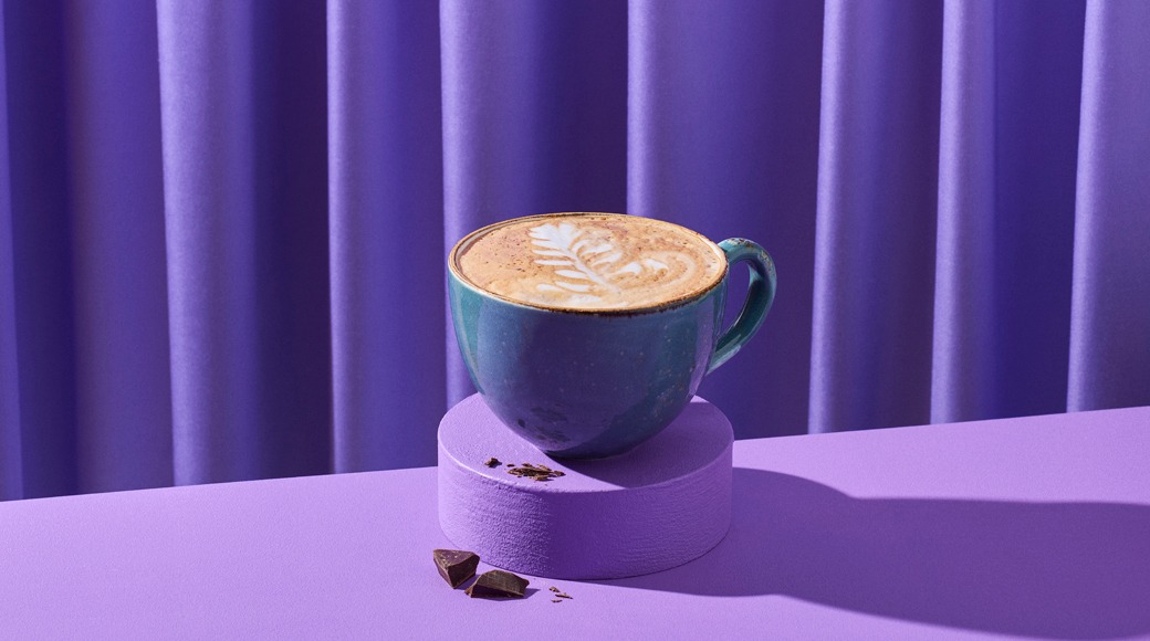 Sinertävä kahvikuppi, jossa on latte-taidekuvio vaahdossa, violetin korokkeen päällä. Vieressä suklaapaloja violettia verhoa vasten.