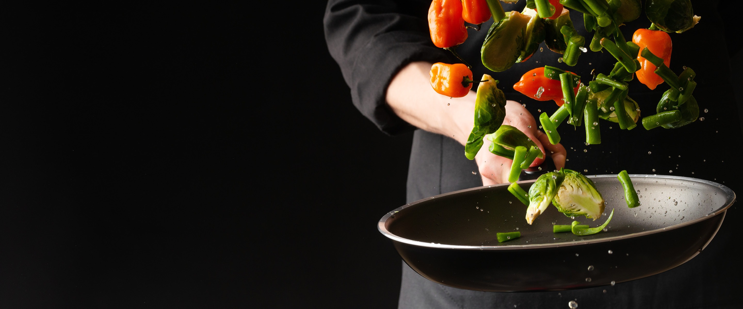 Chef tossing vegetables into a frying pan. Colorful bell peppers, green beans, and brussels sprouts are mid-air with water droplets splashing around.
