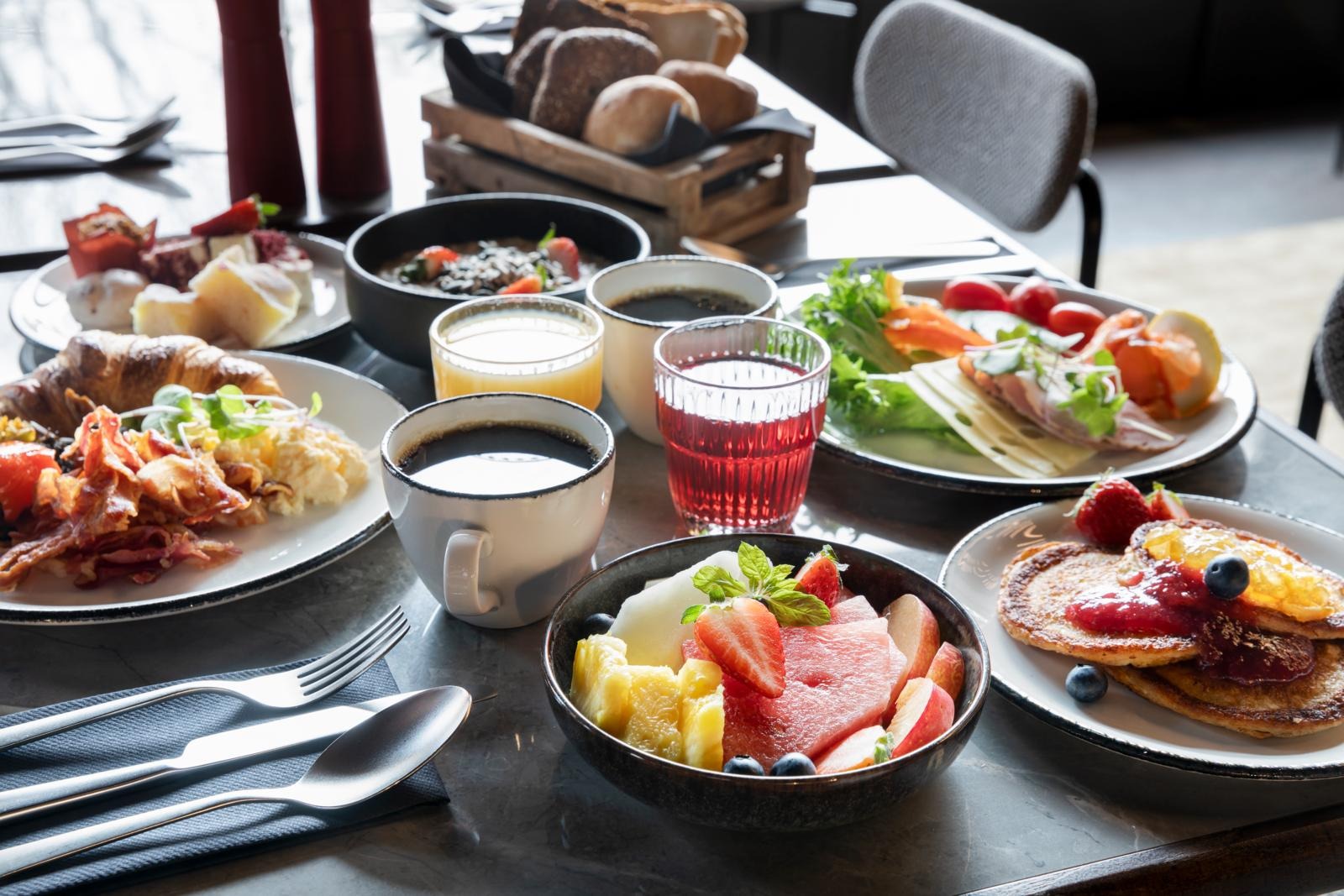 Ett varierat frukostbord med frukt, pannkakor, croissanter, bacon och sallad. Kaffe, juice och en brödkorg kompletterar måltiden.