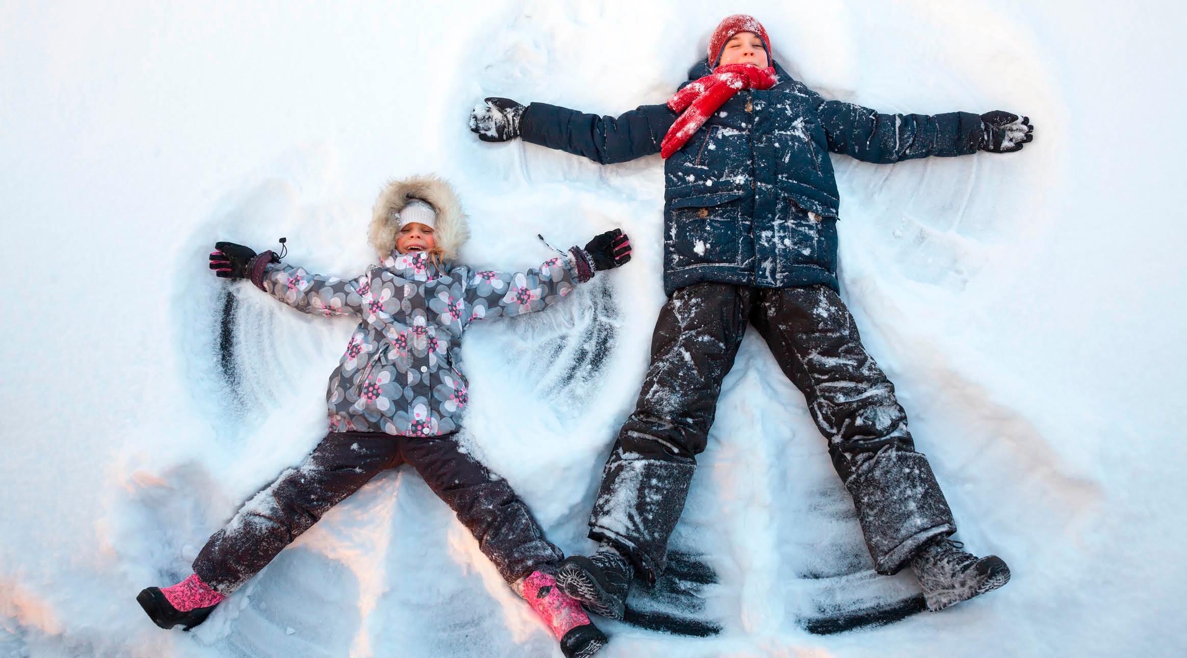 Två barn ligger i snön och gör snöänglar. Ett barn har en blommig jacka och det andra har en blå jacka med röd halsduk.