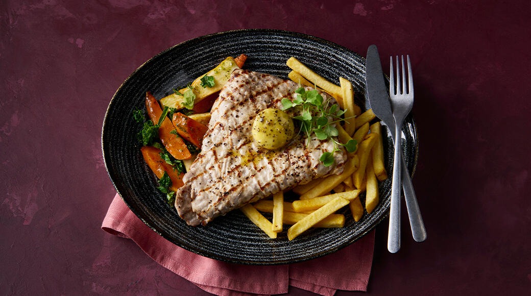 Grilled steak topped with a pat of butter, served with fries and roasted vegetables on a dark plate. A fork and knife lie beside it.