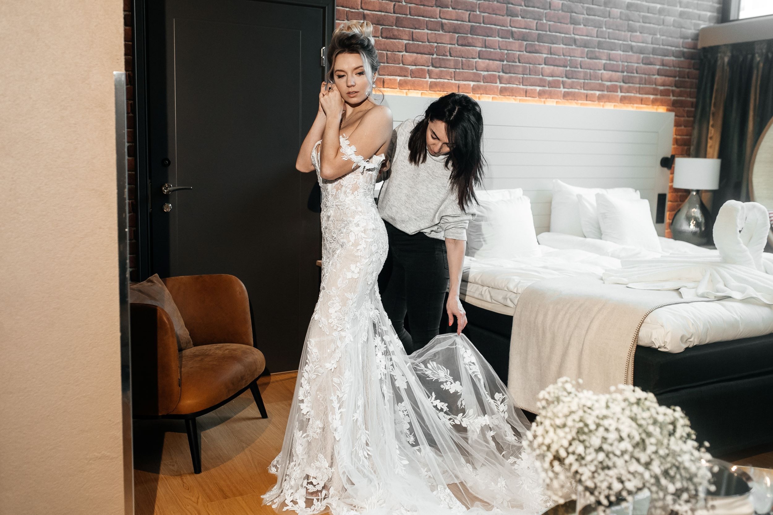 Hotellihuoneessa tapahtuvat hääpukuvalmistelut, toinen henkilö auttaa säätämään pitsisen valkean hääpuvun helmaa.