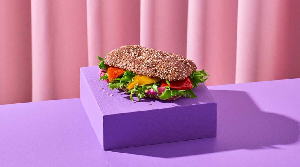 Täysjyväsämpylä, jonka välissä on salaattia, paahdettua paprikaa ja punaista tahnaa, violetilla korokkeella vaaleanpunaista verhoa vasten.