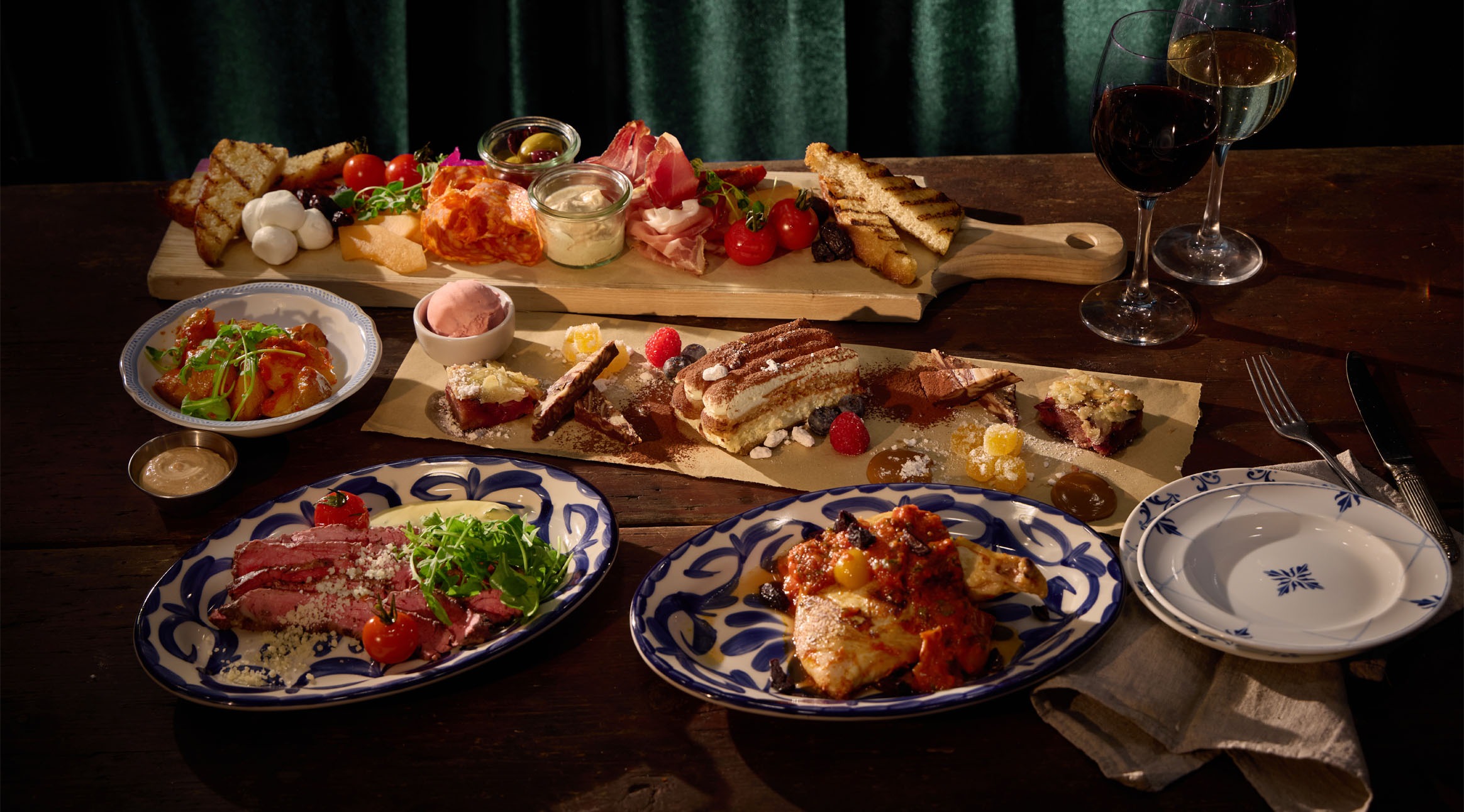 Runsas ateriavalikoima: antipastilautanen, jälkiruoka-assortii, liha-annos, kana-annos ja viinilaseja rustiikilla puupöydällä vihreää taustaa vasten.