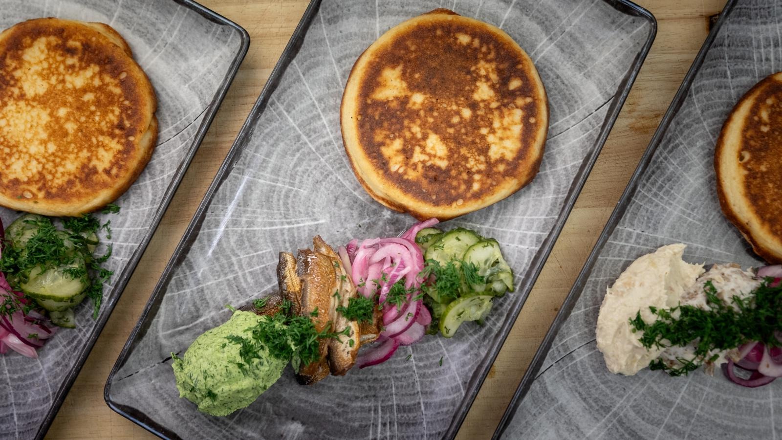 Three plates with fried small pancakes served with various accompaniments: fish, green paste, red onions, cucumbers, and fresh herbs.