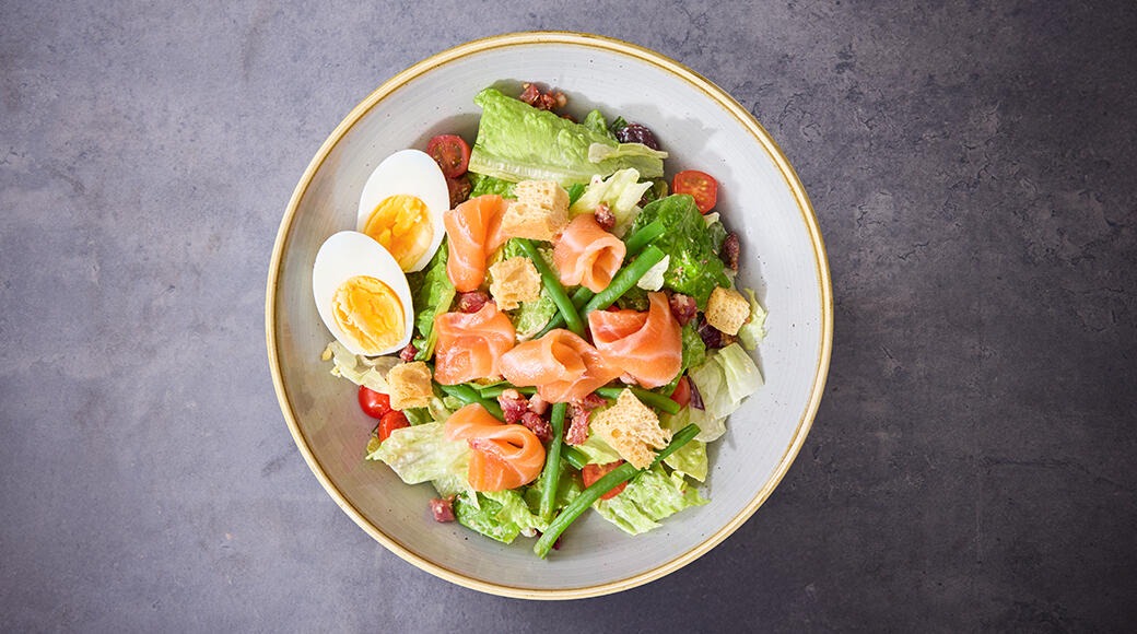 Salaatti, jossa on lohta, halkaisuja keitettyjä munia, vihreää salaattia, tomaatteja, vihreitä papuja ja krutonkeja vaalealla lautasella.